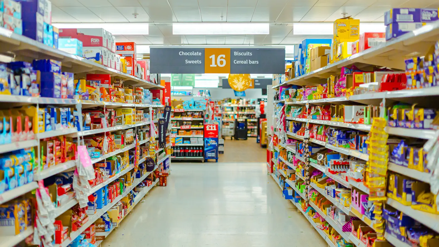 A long, brightly lit supermarket aisle with packed shelves on both sides