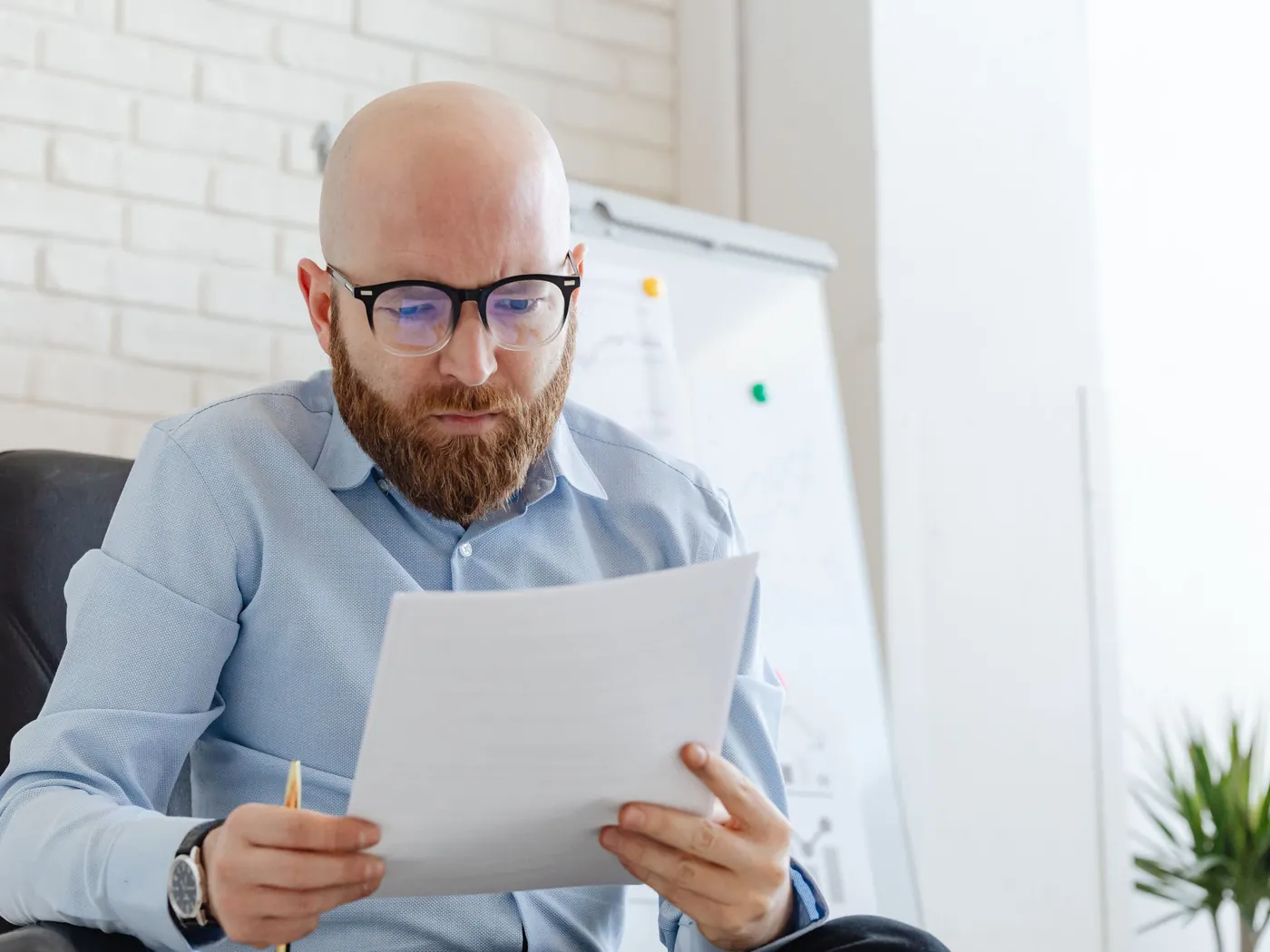 A man in glasses reading a printed document closely at his office desk