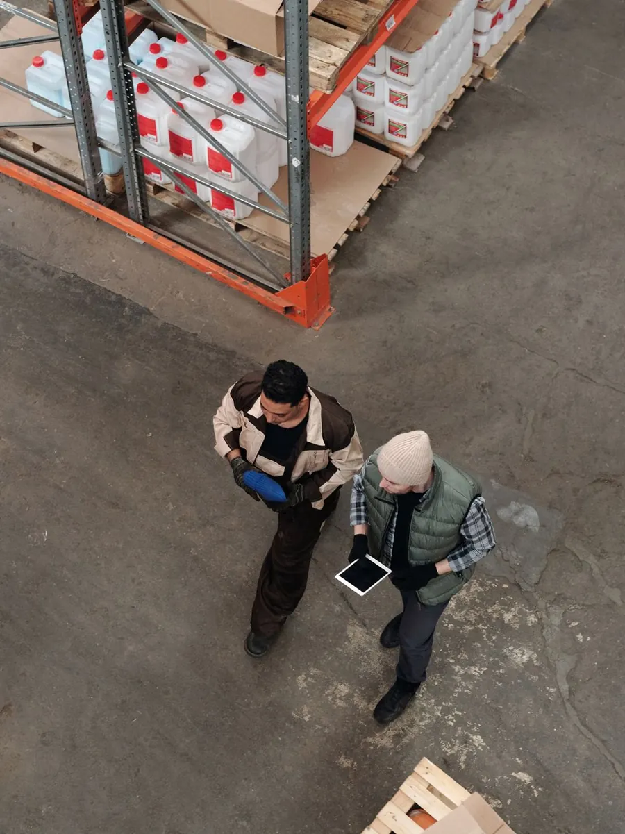 An overhead view of two warehouse workers checking a tablet beside pallets