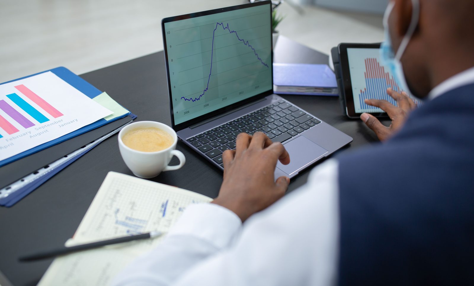 Analyst reading a multi-year forecast line chart on a laptop next to a tablet of bar charts