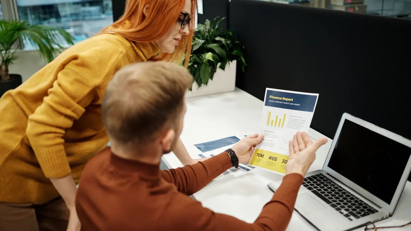 Two colleagues at an office desk going through a printed finance report next to a laptop