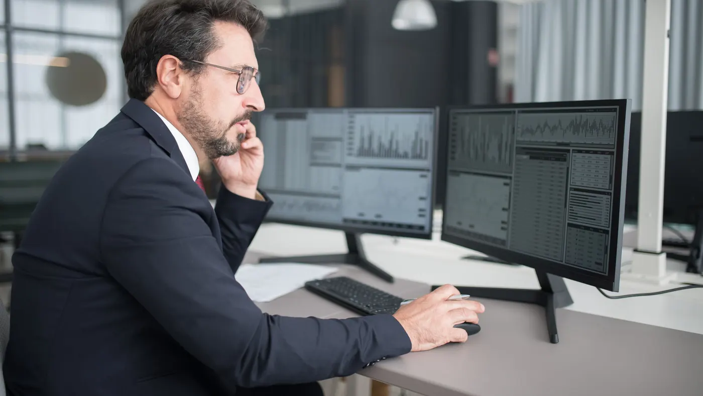 A man on a phone call at his desk, looking at charts on two computer screens