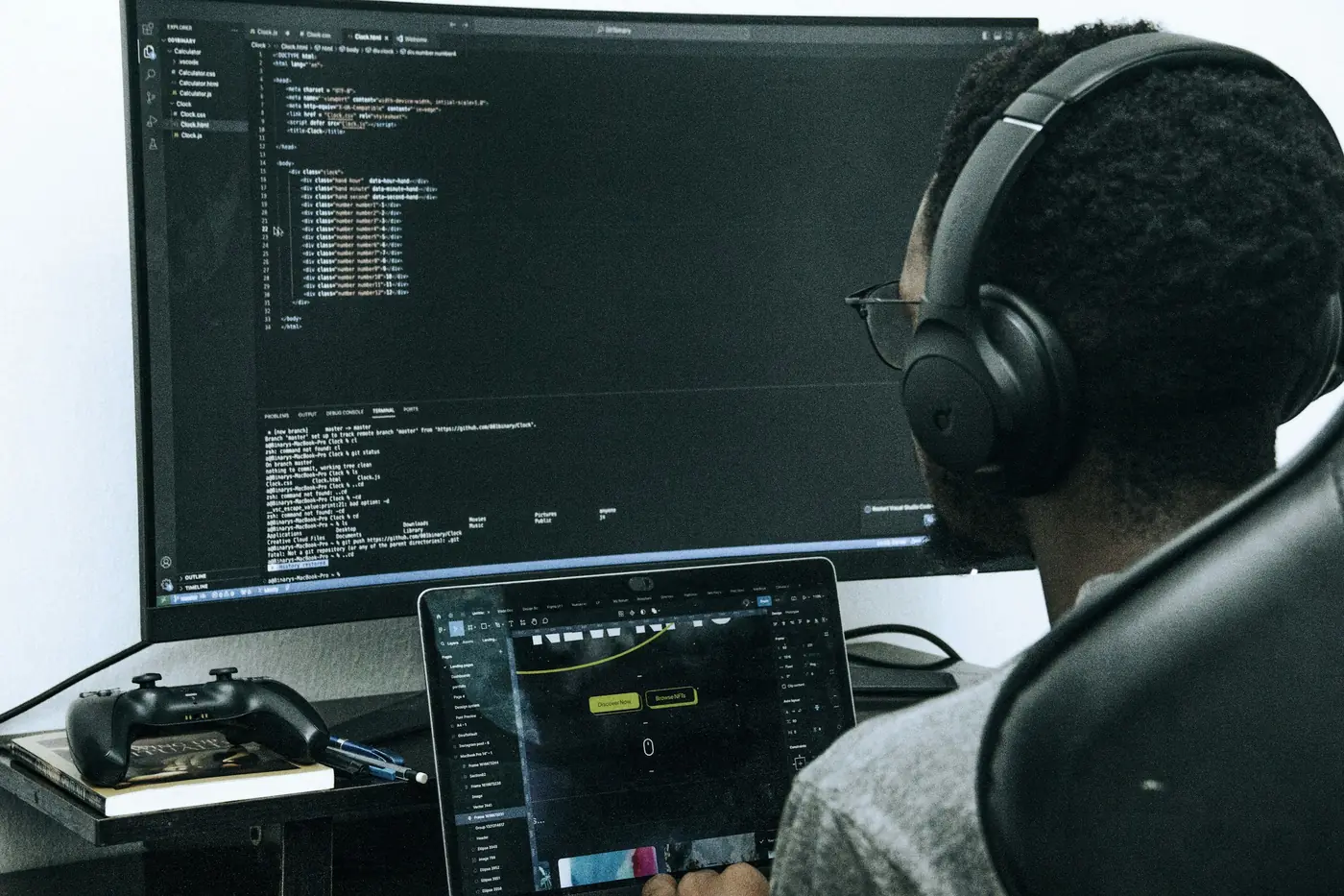 Developer wearing headphones and glasses, seen from behind, working at a large monitor showing HTML code and a laptop with a design tool open