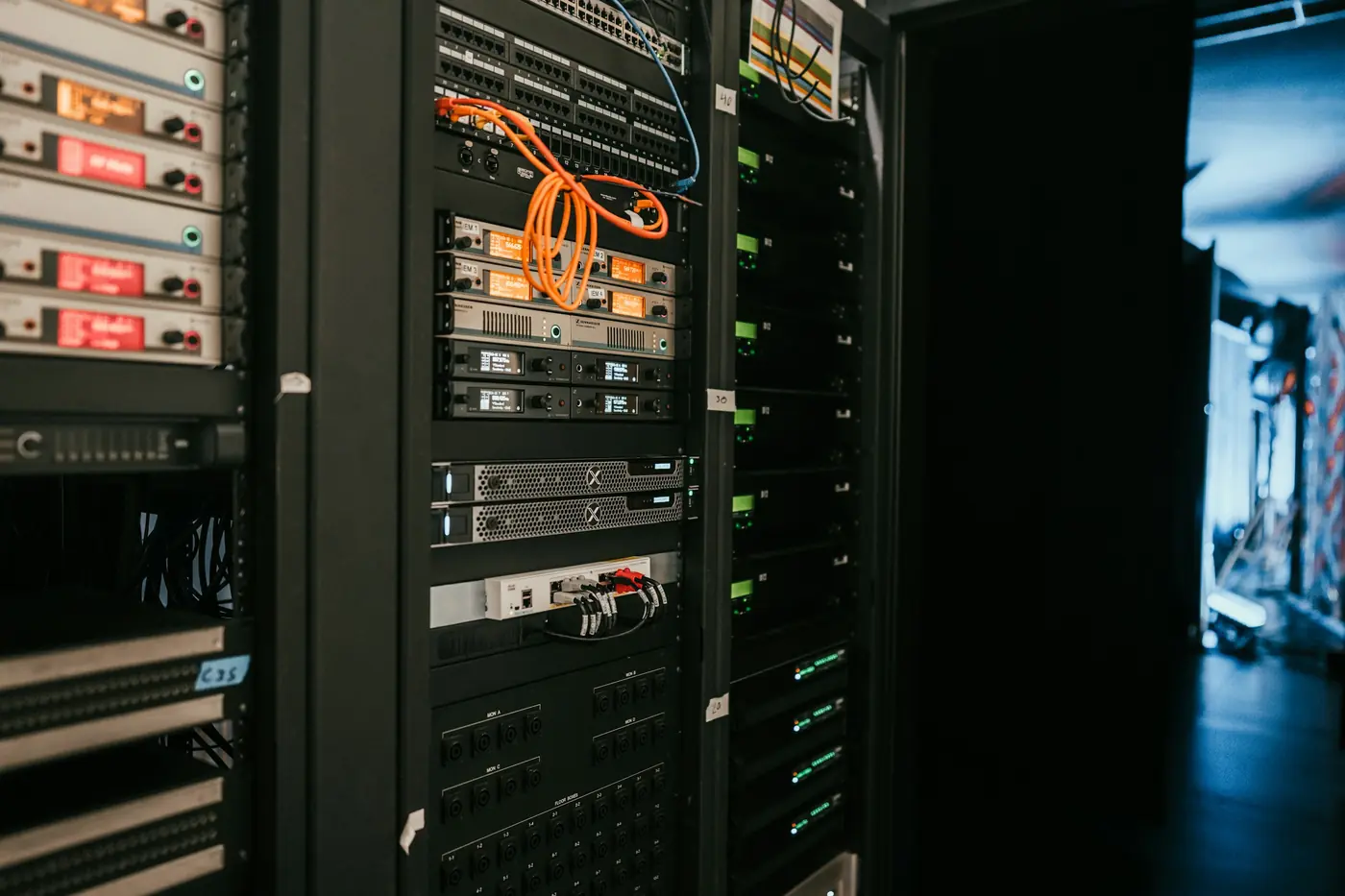 Dark equipment racks filled with patch panels, rack-mounted units with small displays and an orange network cable