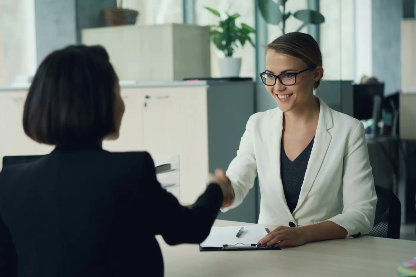 Smiling woman in glasses and a white blazer shaking hands across a desk with a dark-haired woman in a modern office