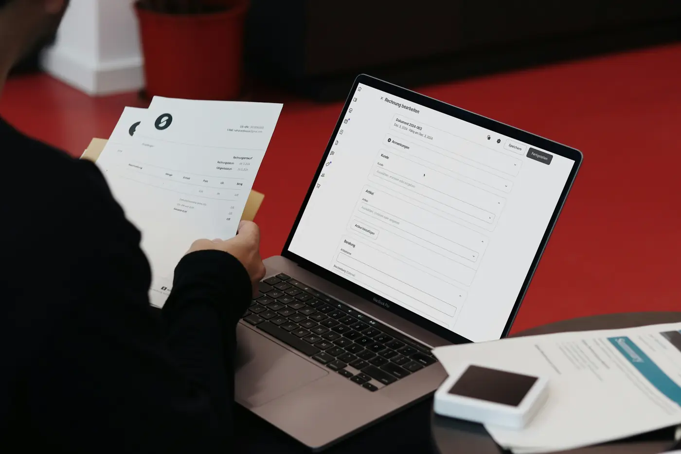 Person holding printed invoices next to a laptop showing an invoice-editing form, on a red table