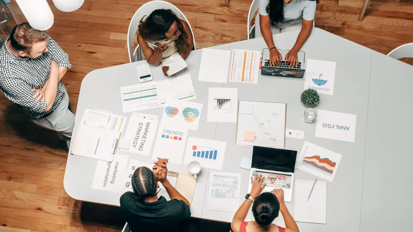 A marketing team seen from above, working around a table covered in charts, notes and laptops